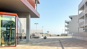 Un edificio moderno con fachada de vidrio, entrada enmarcada en rojo y un paseo empedrado que conduce al frente marítimo.