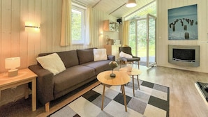Una sala de estar acogedora con un sofá, una mesita redonda y un televisor. Hay una ventana con cortinas, una puerta que da al exterior y un cuadro en la pared.