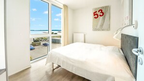 Une chambre d’hôtel avec un grand lit, une vue sur la mer par une porte coulissante, le numéro « 53 » sur le mur et une lampe moderne.