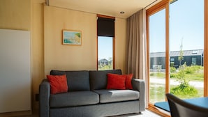 Una habitación moderna con un sofá gris, cojines rojos, un cuadro en la pared y vistas a un edificio y zonas verdes.