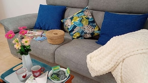Ein graues Sofa mit blauen und blumigen Kissen, einer weißen Decke, einem gewebten Korb, einer Vase mit Blumen, einer Ablage mit Tassen und einer Schüssel mit Snacks sowie eine Zeitschrift.