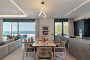 A modern dining area with a large table, chairs, and a view of the ocean. - Nelisa, A Private Sanctuary of Sun, Sea & Timeless Luxury in Crete (Vlychada)