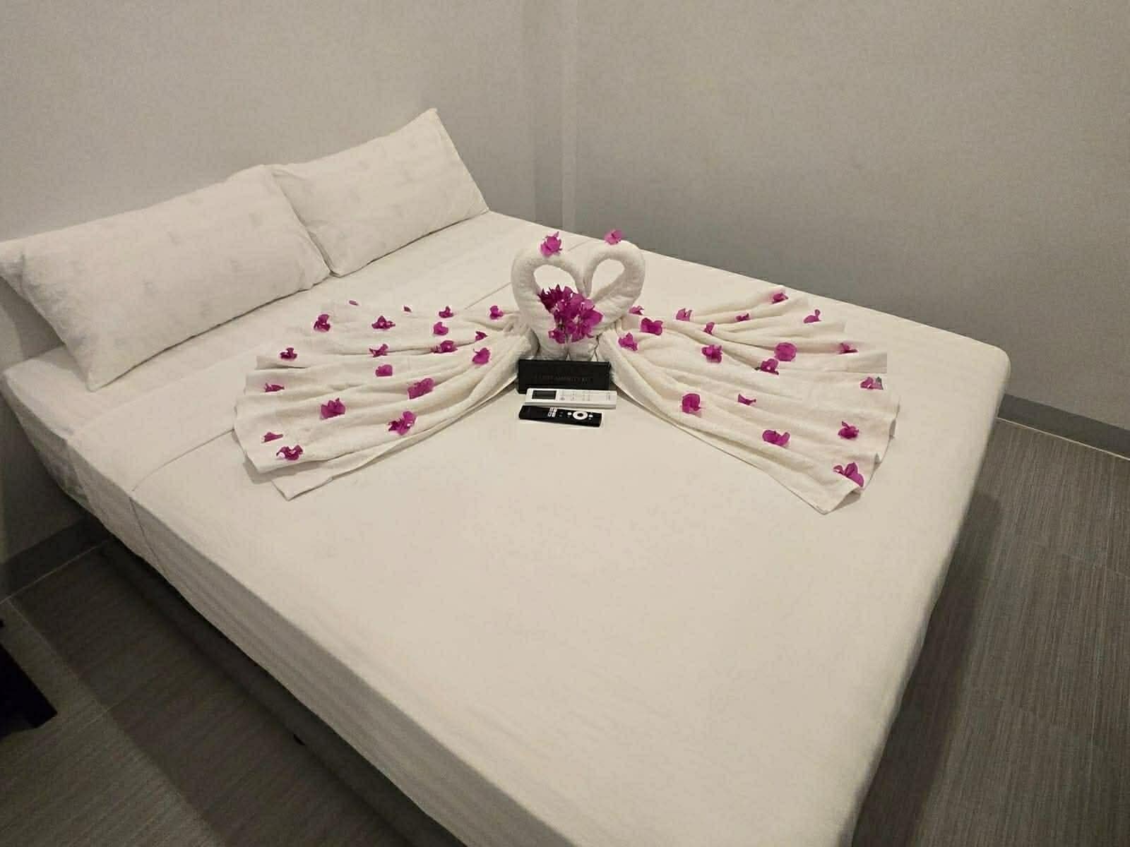 A neatly made bed with a towel arranged in a swan shape, adorned with pink flowers, and a black object on the bed.