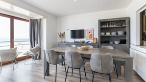 A modern kitchen with a dining area, featuring a wooden table, grey chairs, a TV, and a view of the sea through large windows. - 3 bedroom accommodation in Nieuwpoort (Nieuwpoort)