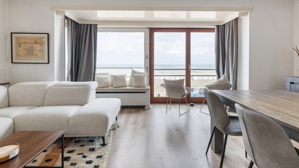 A modern living room with a sofa, a dining table, and chairs, a large window with a view of the beach, and a framed picture on the wall. - 3 bedroom accommodation in Nieuwpoort (Nieuwpoort)