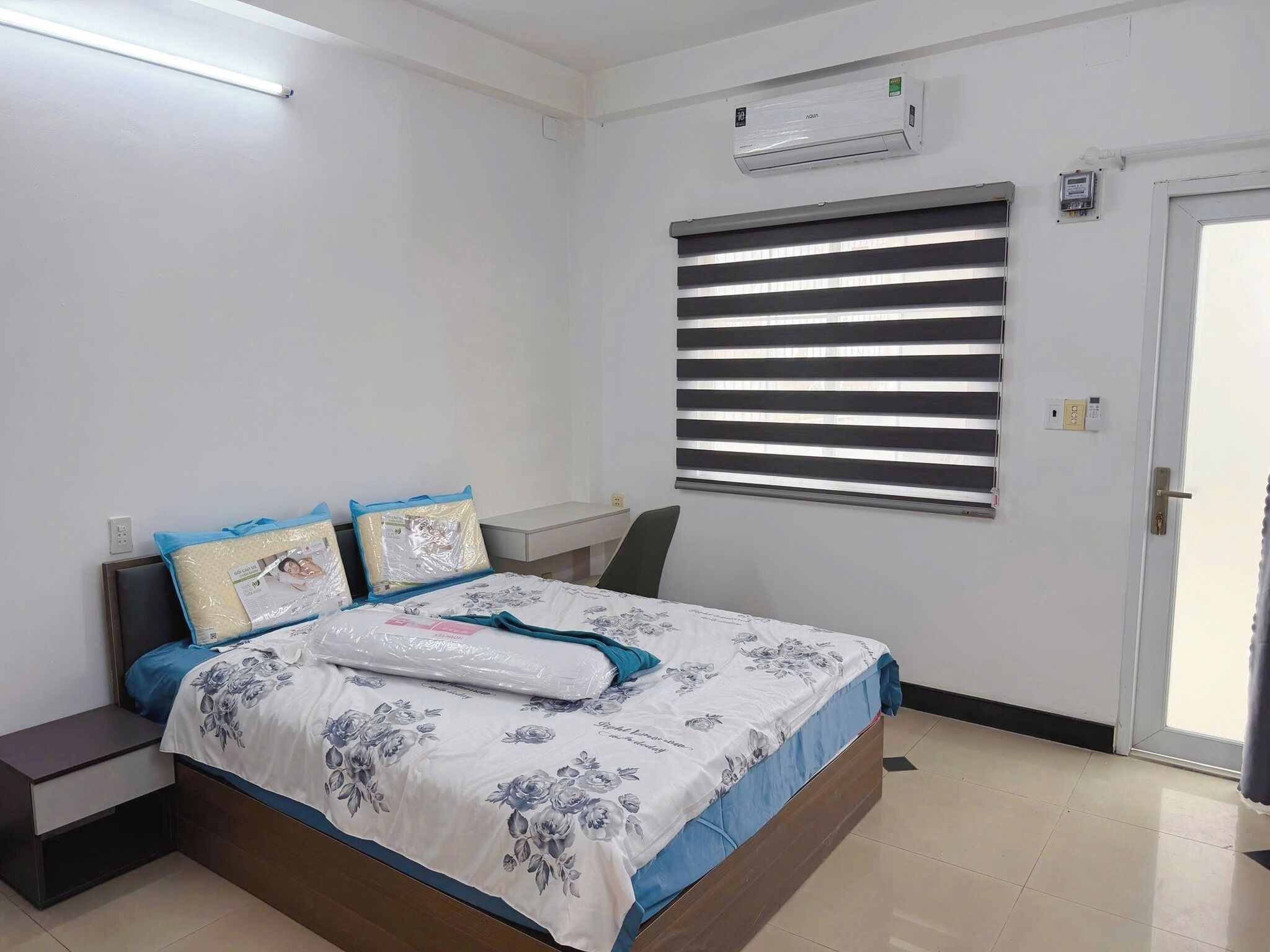 Um quarto com cama, mesas de cabeceira, ar condicionado, janela com persianas e uma porta.