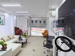A modern living room with a sofa, coffee table, and chairs, a large window with a view of the street, and a mounted TV.