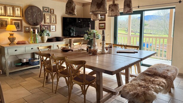 A dining area with a wooden table, chairs, and a bench. There are hanging lights, a clock, and a TV on the wall.