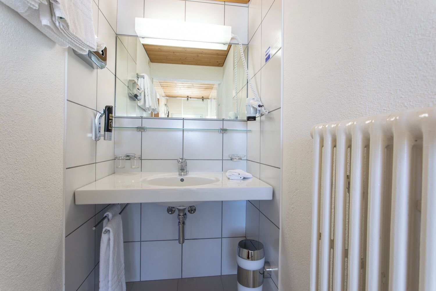 Un baño moderno con un lavabo blanco, espejo y paredes con azulejos. Se observa un perchero, cabina de ducha de vidrio y un radiador.