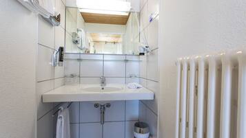 Un baño moderno con un lavabo blanco, espejo y paredes con azulejos. Se observa un perchero, cabina de ducha de vidrio y un radiador.
