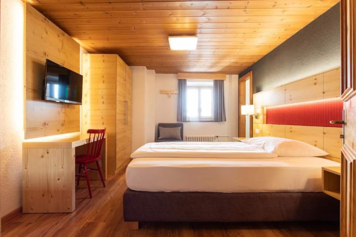 Ein Zimmer mit Holzboden, einem Bett mit weißer Bettwäsche, einem roten Sessel, einem an der Wand montierten Fernseher und einem Fenster mit Vorhängen.