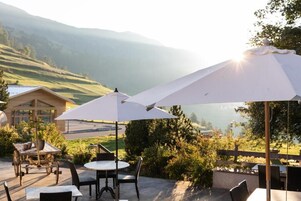 Un patio al aire libre con mesas y sillas, sombrillas blancas y vistas a un paisaje montañoso.