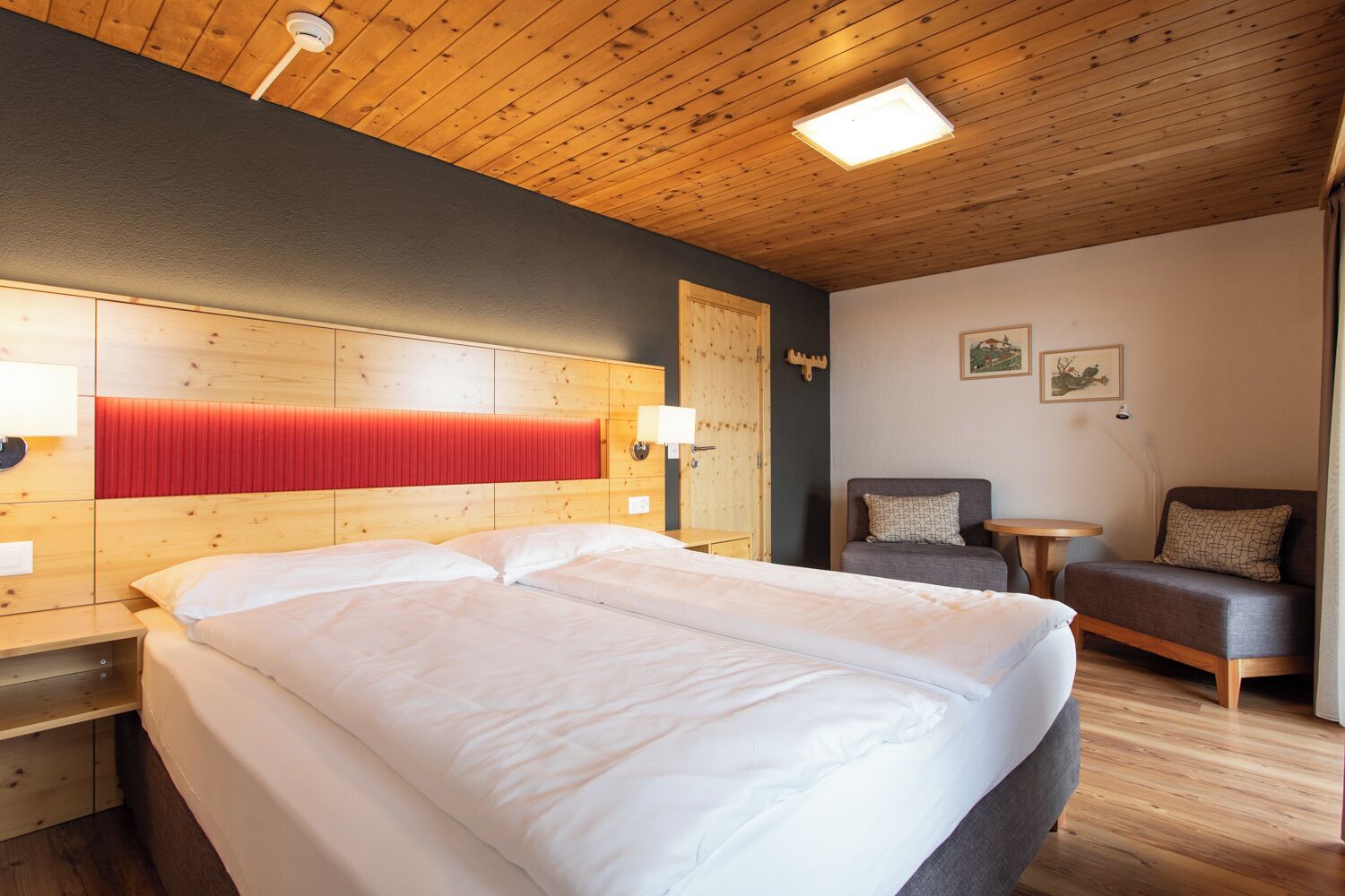 A hotel room with a large bed, a wooden headboard, a red accent panel, a small table, a sofa, and a wall-mounted lamp.
