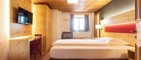A hotel room with a wooden ceiling, a bed with white linens, a red chair, a TV mounted on the wall, and a window with curtains.