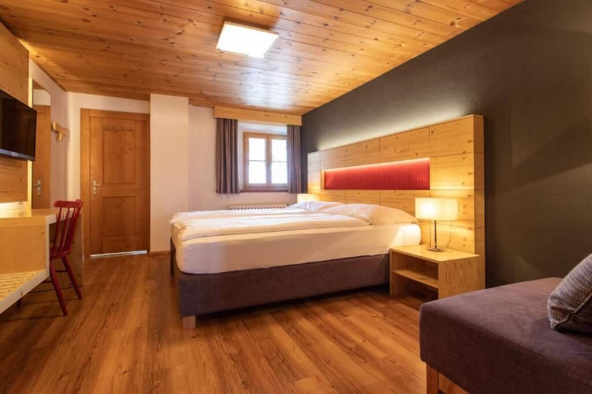 Ein Hotelzimmer mit Holzboden, einem Bett mit rotem Kopfteil, einem Nachttisch mit Lampe, einem Sessel und einem an der Wand montierten Fernseher.