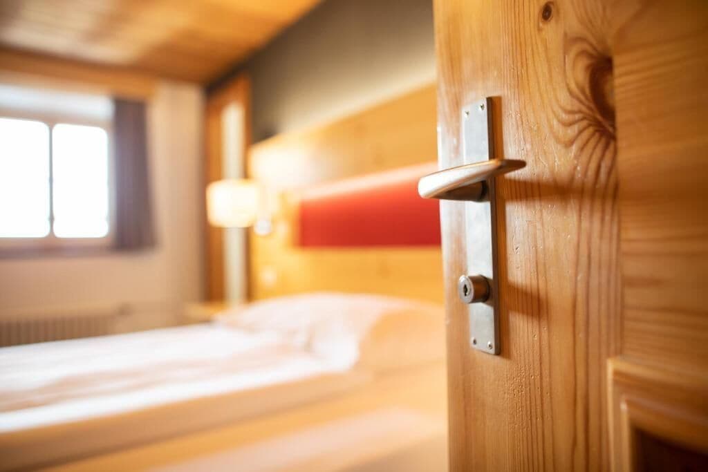A wooden door with a metal handle and a lock, part of a room with a bed and a window.