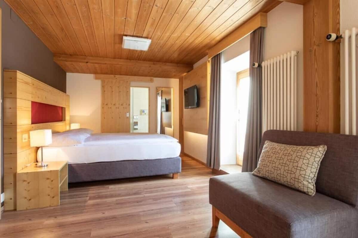 Ein Hotelzimmer mit Holzboden, einem Bett mit grauer Matratze, einem braunen Sofa, einem an der Wand montierten Fernseher und einem Fenster mit Vorhängen.
