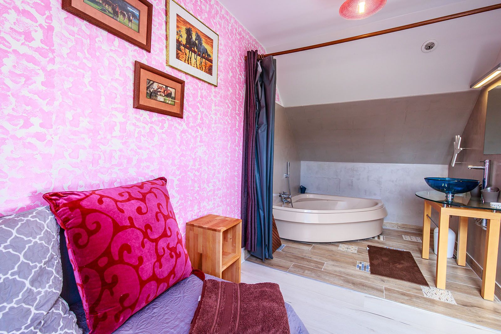 Een slaapkamer met een bed, nachtkastje, houten bijzettafeltje, groot raam met gordijnen, en een badkamer met badkuip, wastafel en spiegel.