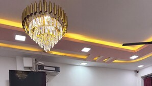 A room with a chandelier, a flat-screen TV, a calendar, and plastic chairs. - The Vow Cartel Ananta (Varanasi)