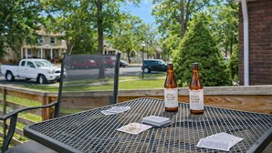 Metallinen pöytä, jonka päällä on kaksi pulloa Honey Orange Tripel -olutta, pakka kortteja ja pieni astia. Pöytä on parvekkeella, jolta avautuu näkymä asuinalueen kadulle, jossa on taloja ja parkkeerattuja autoja.
