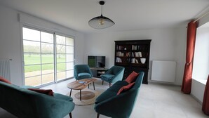 Un salón amplio con un gran ventanal corredera, una estantería negra, sillones azul verdosos, una mesa de centro y una lámpara colgante.