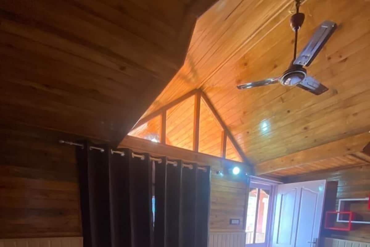 A wooden cabin bedroom with a bed, curtains, a ceiling fan, and a view of the outside.