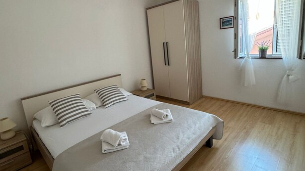 A bedroom with a large bed, a wardrobe, a bedside table, and a window with curtains. - Beautiful Apartment Bodulo 1 (Povljana)