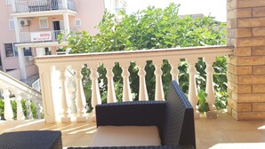 A balcony with wicker furniture, a white balustrade, and a building with balconies and greenery in the background. - Beautiful Apartment Bodulo 1 (Povljana)