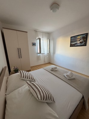 Ein ordentlich eingerichtetes Schlafzimmer mit einem Bett, Kissen, einem Nachttisch mit Lampe und Pflanze, einem Kleiderschrank und einem Fenster mit Vorhängen.
