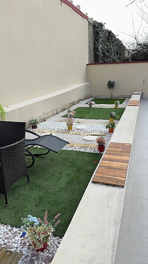 A patio area with a black outdoor sofa, a small table, and a variety of potted plants. There is a pathway with stepping stones leading to a wooden deck. - Royal Sapphire Apartment With Royal Garden (Le Bourget)