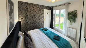 A bedroom with a bed, a wardrobe, a window with a view of the garden, and a wall with a decorative pattern. - Royal Sapphire Apartment With Royal Garden (Le Bourget)