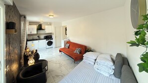 A compact living space with a bed, a sofa, a small table, and a kitchen area in the background. - Royal Sapphire Apartment With Royal Garden (Le Bourget)