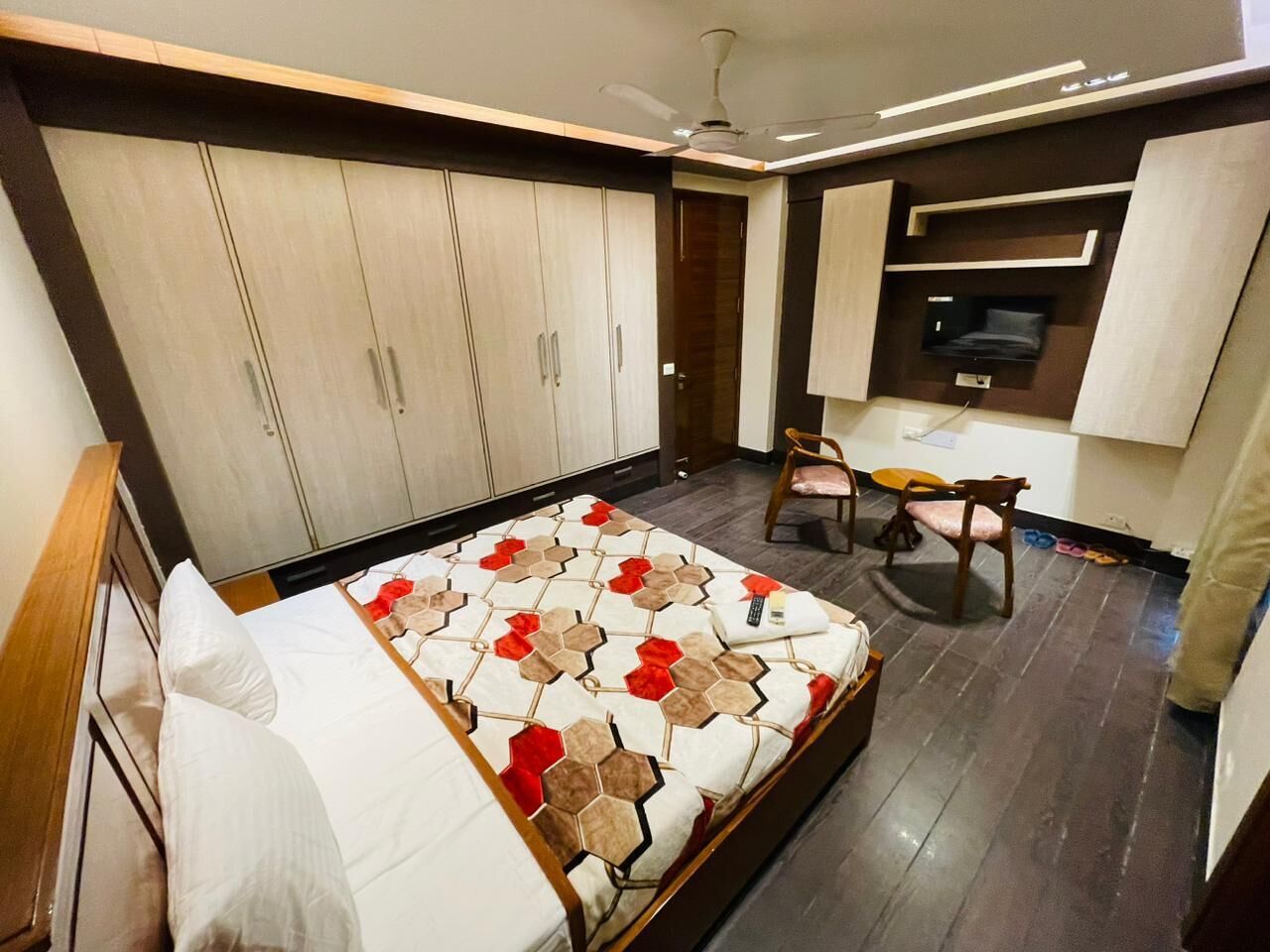 A bedroom with a bed, a desk, and a chair. There is a wardrobe and a television on the wall.