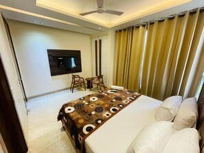 A hotel room with a bed, a TV mounted on the wall, a desk with chairs, and a ceiling fan. - Fortune Home Service Apartment 4Bhk D163 (New Delhi)