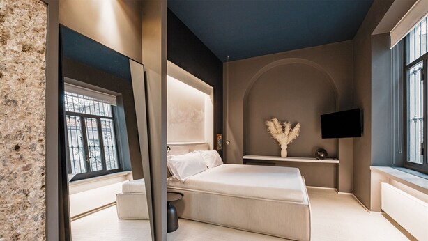 A modern bedroom with a large bed, a bedside table, a small seating area, and a TV mounted on the wall. - ARCHILOFT Brera Gae Aulenti Moscova (Milano)