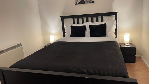 Una cama bien hecha con cabecera negra, dos lámparas de noche y un cuadro colgado en la pared.