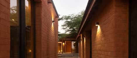 A corridor with brick walls and large windows, leading to a covered area with doors.