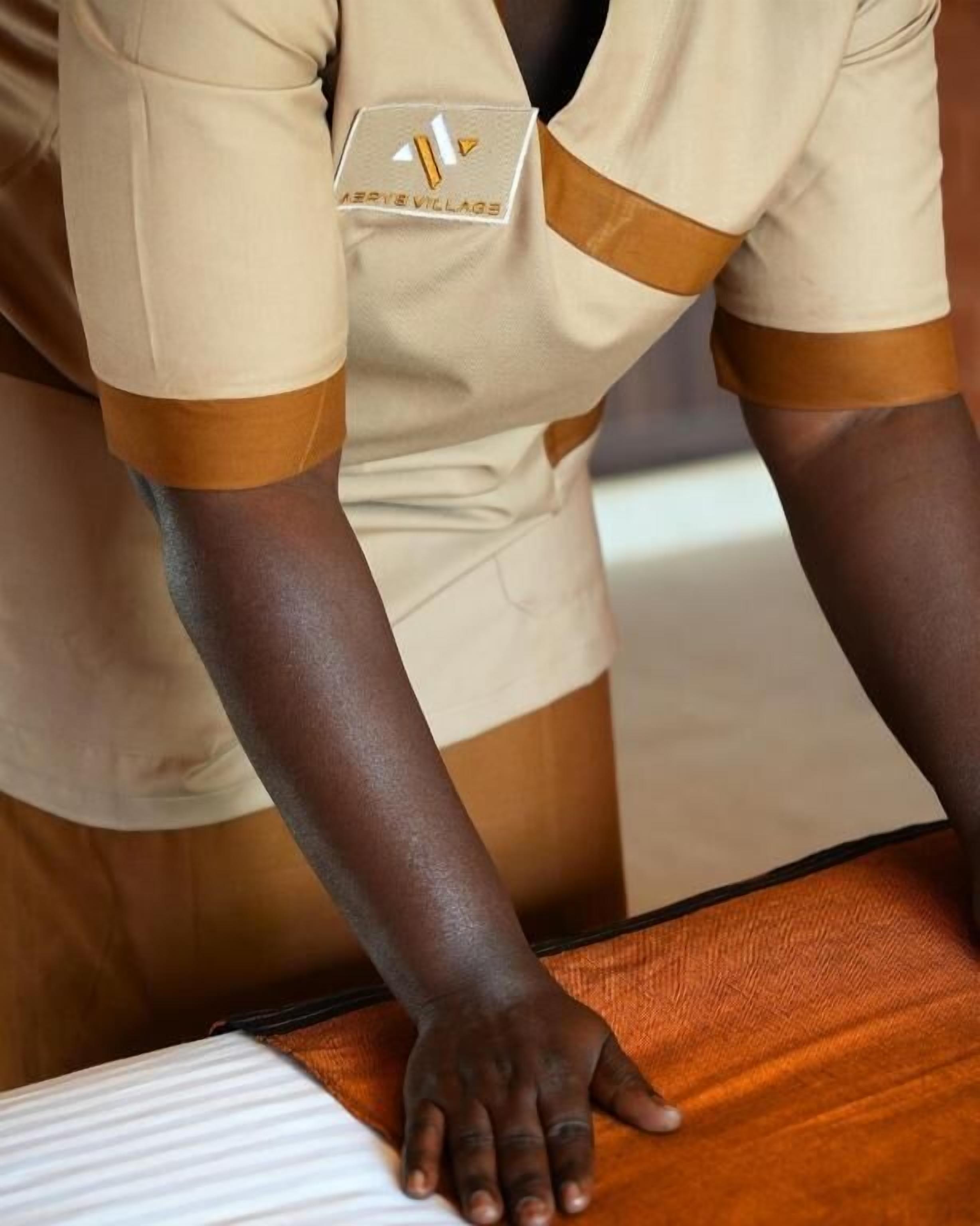 A person in a beige uniform with a name tag, seated at a wooden table with a white-striped tablecloth.