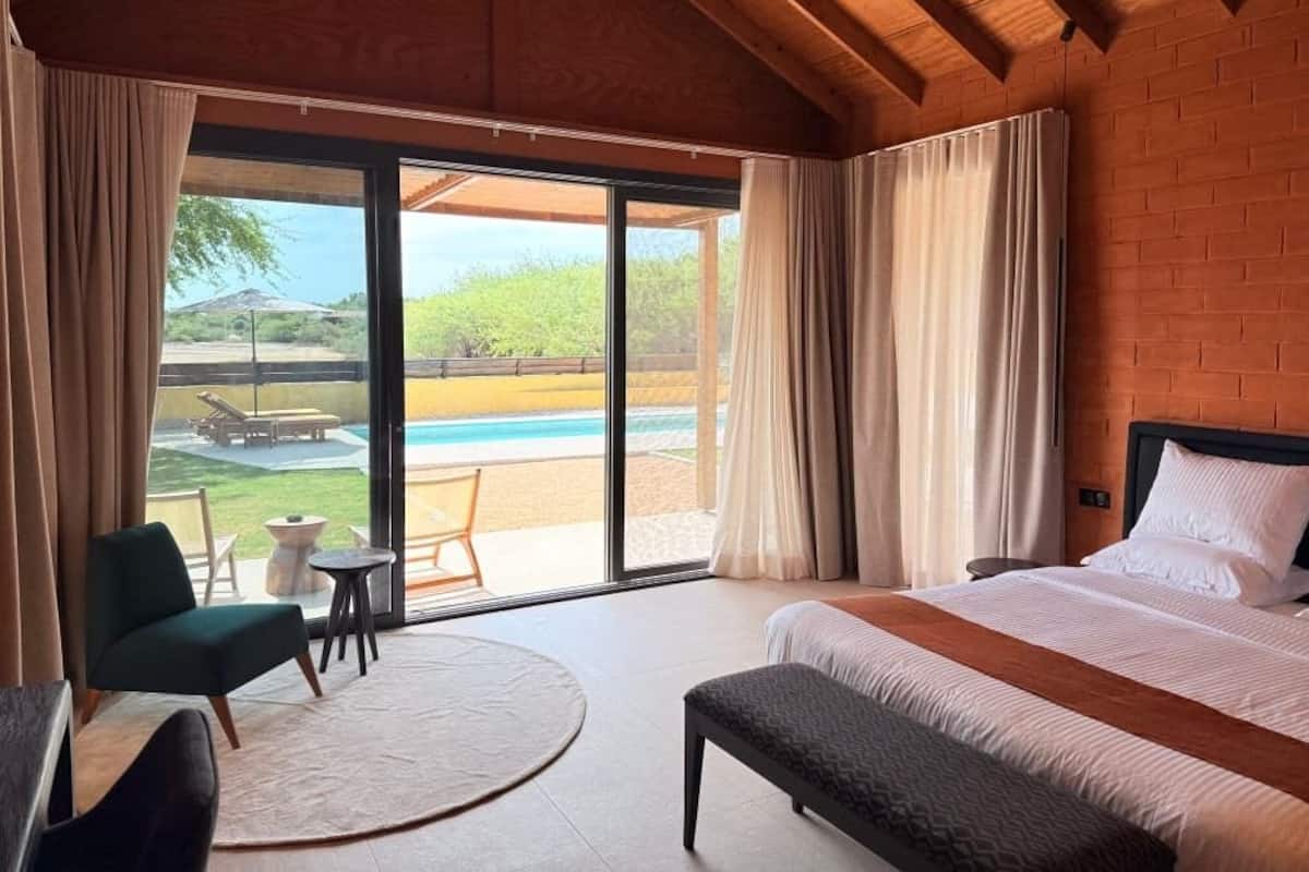 A bedroom with a large bed, a bench, a chair, a small table, and a view of an outdoor pool and garden.