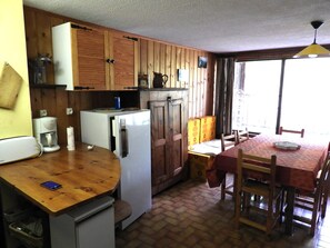 A compact kitchen with wooden cabinets, a refrigerator, a coffee maker, and a dining table with chairs. - Family studio facing the forest at the start of hiking and cross-country skiing trails (Molines en Queyras)