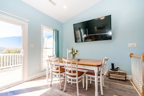 A dining area with a wooden table and white chairs, a flat-screen TV mounted on the wall, and a view through a sliding glass door. - Shore Thing - 4BR, Dog Friendly, Community Pool, Ocean Views (Surf City)