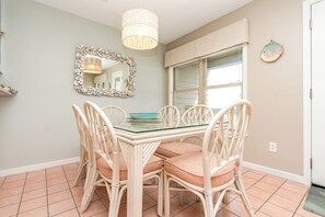 A dining area with a glass table surrounded by wicker chairs, a decorative mirror, and a window with blinds. - Serenity Hearts - Oceanfront Serenity Point Home - 2BR! (Topsail Beach)