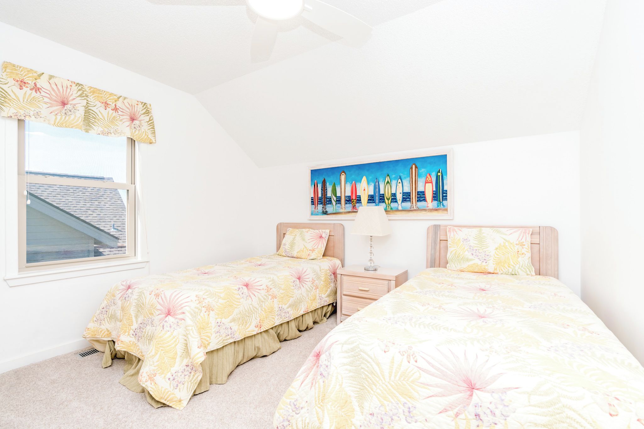 A bedroom with two beds, a nightstand, a lamp, a window with a view of the roof, and a painting of surfboards.