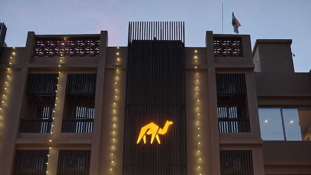 A hotel entrance with a camel logo, large illuminated sign, and a reception area visible through glass doors. - Hotel Camel managed by CLAY (Mundra)