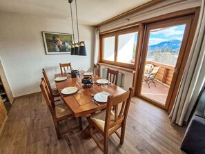 Un comedor con una mesa de madera dispuesta para cuatro, sillas, un cuadro en la pared y vistas a través de un ventanal.