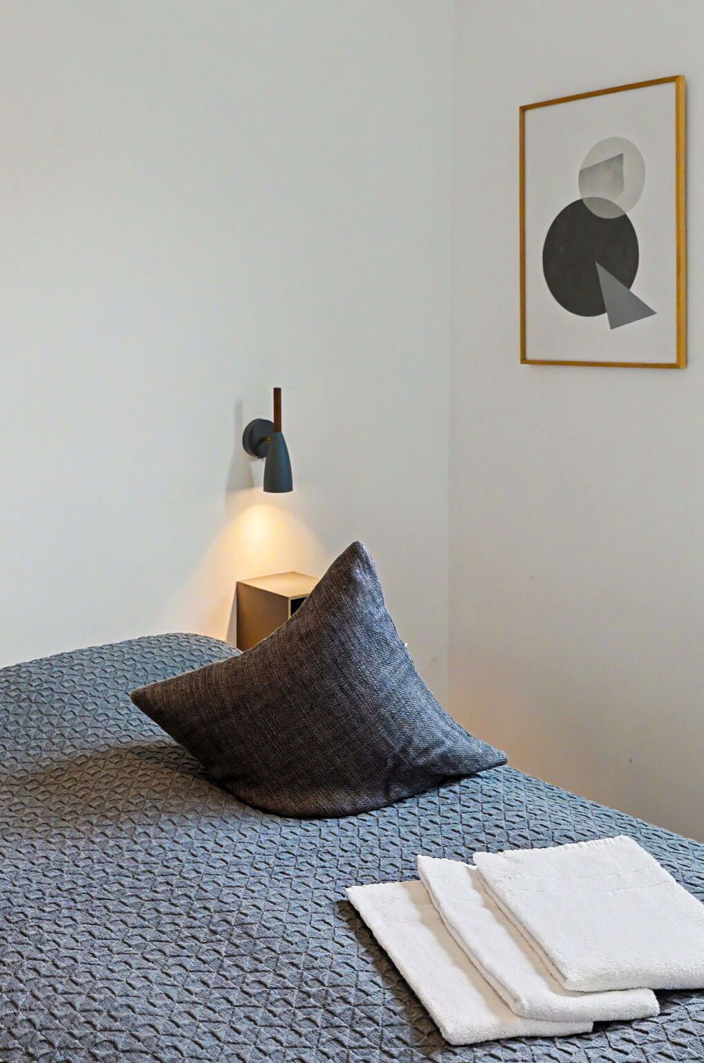 Ein ordentlich bezogenes Bett mit einer gemusterten grauen Decke, einem einzelnen grauen Kopfkissen und einem gefalteten weißen Handtuch.