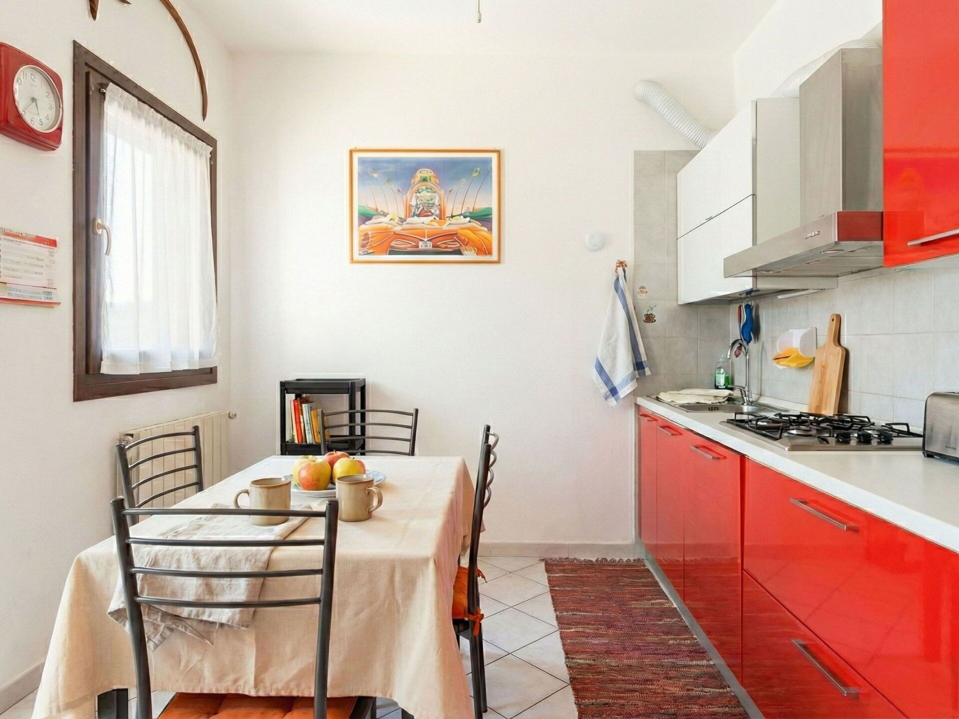Une cuisine de taille réduite avec des placards rouges, une table à manger avec des chaises, une horloge et un tableau encadré au mur.