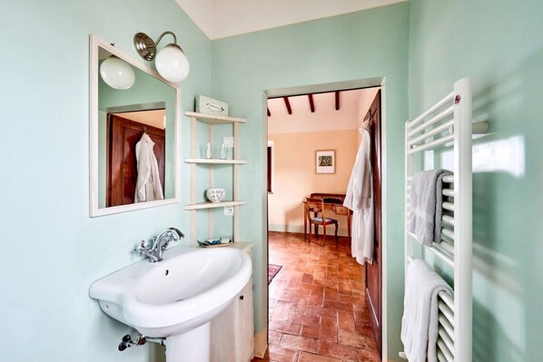 Un baño con lavamanos, espejo y estanterías. Se ve una puerta que da a un comedor con una silla y una mesa.