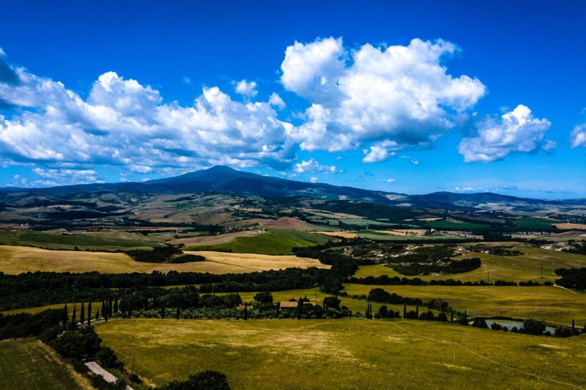 A panoramic view of a rural landscape with rolling hills, a mountain in the distance, and a clear blue sky with scattered clouds.