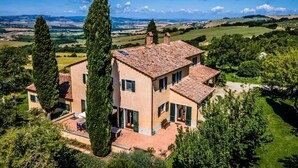 Une maison à deux étages, avec un toit recouvert de tuiles, entourée de verdure et avec des collines en arrière-plan.